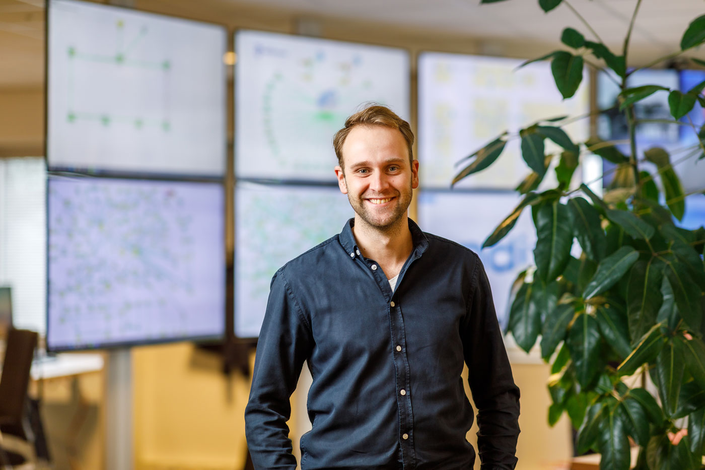Man smiling in office with large green plant and multiple computer monitors displaying data in the background.