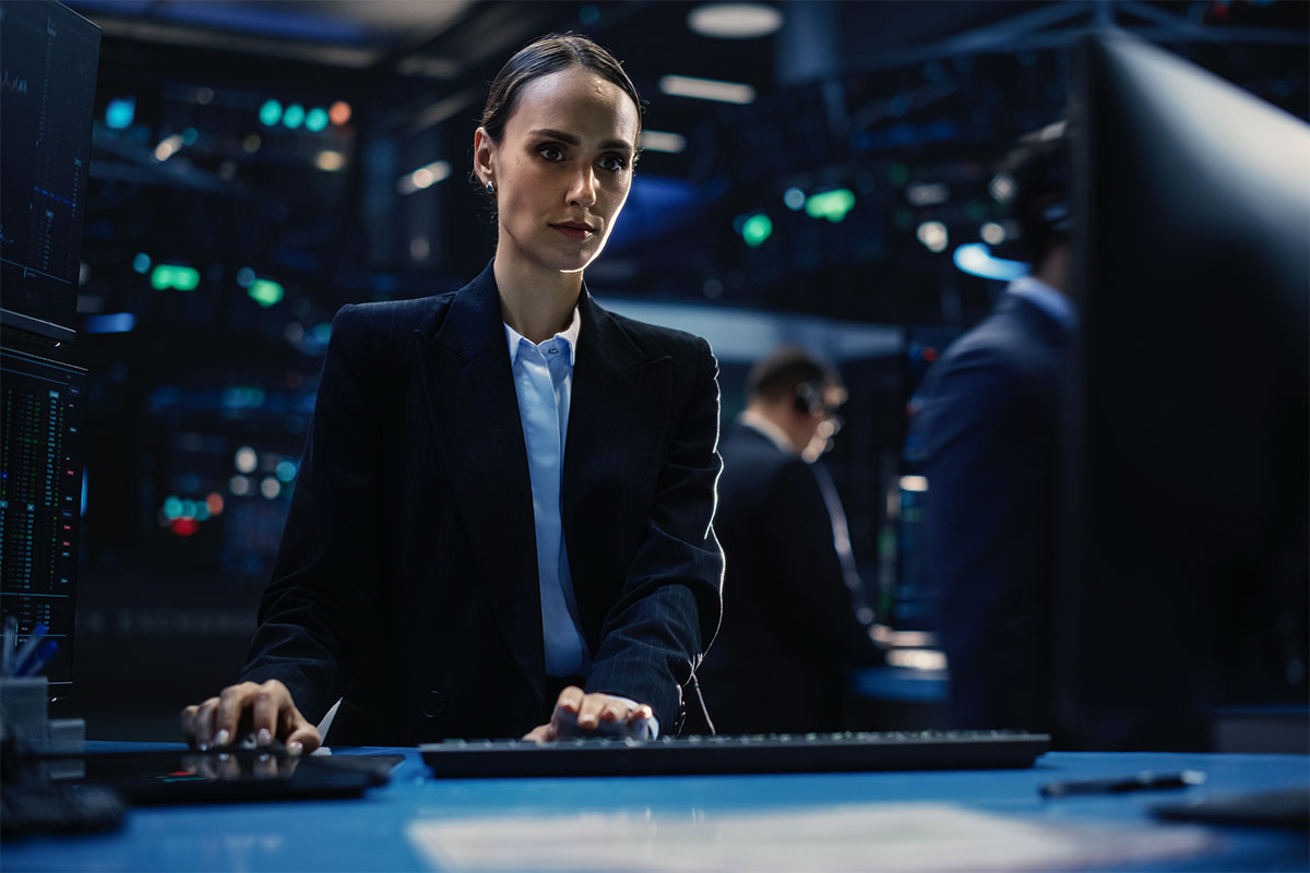 Woman in a suit working at a computer in a dimly lit office with monitors and people in the background.
