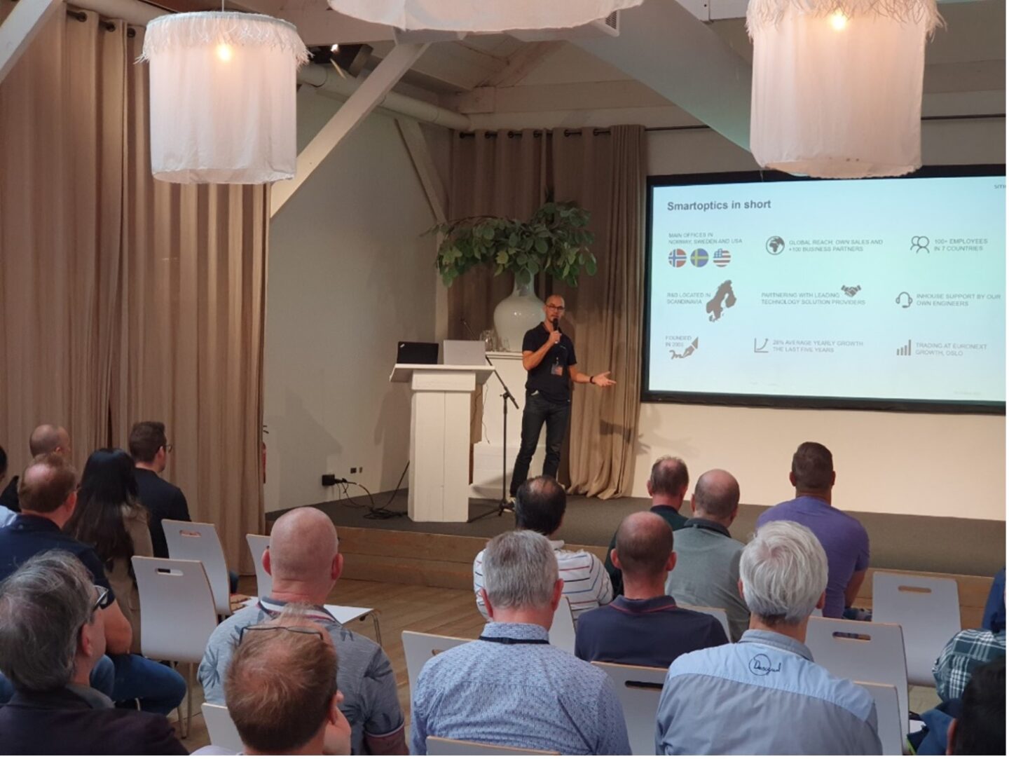A man gives a presentation to an audience in a room, with a slide about Smartoptics shown on a screen.