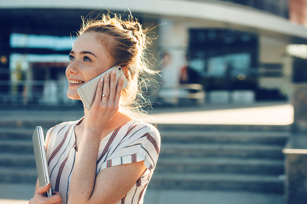 Smiling woman holding a phone to her ear and carrying a laptop, standing outdoors in sunlight.